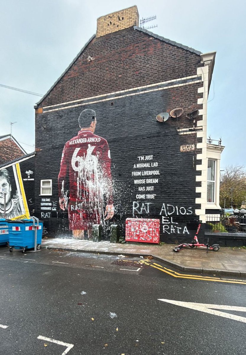 Large mural on brick wall of terraced house depicts Trent Alexander-Arnold in Liverpool FC red jersey with number 6, posed spraying champagne from bottle, dynamic action pose with jersey appearing wet from spray, text graffiti nearby reads RAT ALEXANDER ARNOLD FROM LIVERPOOL YOU ARE A RAT, urban street setting with blue bin, red box, scooter, and adjacent wall art.