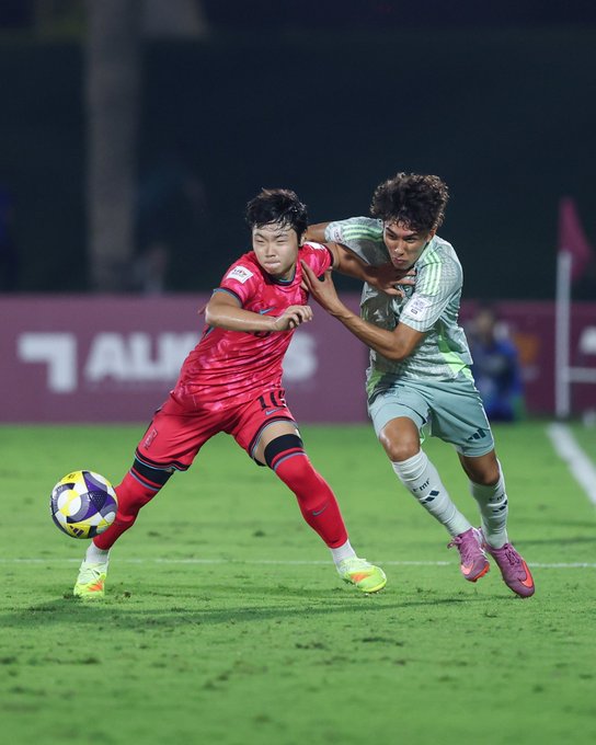 Two young male soccer players in action on a green field during a night match under stadium lights one in a red jersey with number 18 marked by a sponsor logo pushing against the other in a green jersey both wearing shorts knee socks and cleats with a yellow and purple soccer ball at their feet red banners in the background.