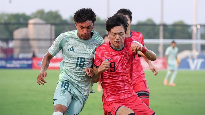 Three soccer players in action on a green field during a match, one in green jersey number 18 pulling the arm of a player in red jersey number 9, another in red jersey behind, stadium banners and sunset sky in background.