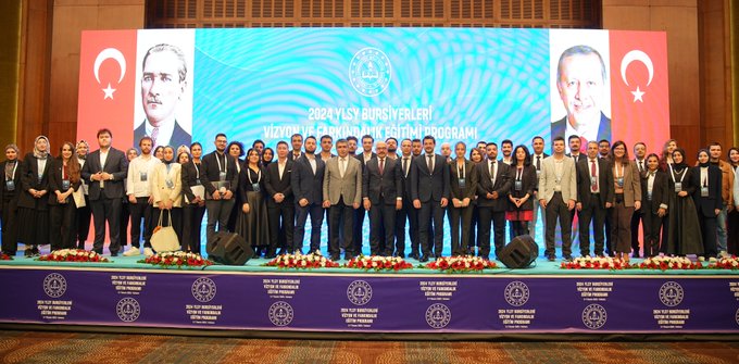 İlk görüntüde bir adam kürsüde konuşma yapıyor, arkasında Nevşehir Hacı Bektaş Veli Üniversitesi logosu ve 2024 YLSY Bursiyerleri Vizyon ve Farkındalık Eğitimi Programı yazısı var, kürsüde Semih Aktekin ve Ankara 3-7 Kasım yazıları görülüyor. İkinci görüntüde mavi arka planda Türk bayrakları ve 2024 YLSY Vizyon ve Farkındalık Programı yazısı, sahne önünde oturmuş bir grup insan ve konuşmacılar var. Üçüncü görüntüde geniş bir salonda mavi ekranlı sahne, Türk bayrakları ve kalabalık bir dinleyici grubu koltuklarda oturuyor. Dördüncü görüntüde grup fotoğrafı, çeşitli kıyafetli erkek ve kadınlar sahnede poz veriyor, arka planda mavi ekran ve Türk bayrakları ile program yazısı.