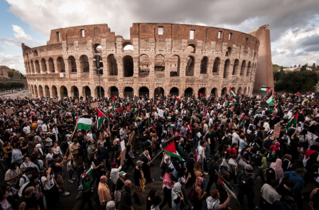 Large crowd of diverse people gathered in front of the ancient Colosseum structure under a cloudy sky, many holding and waving Palestinian flags with green white red and black colors, some wearing keffiyehs, standing on paved ground with barriers visible.