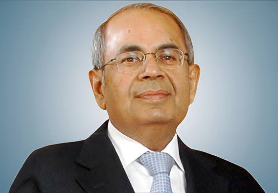 A formal portrait of an older Indian man with short gray hair, wearing eyeglasses, a dark suit jacket, white shirt, and blue tie, smiling slightly against a blue background.