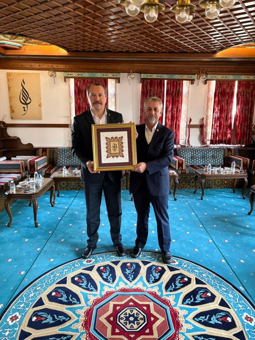 First image shows two men in dark suits standing in a traditional room with wooden ceiling, red curtains, blue carpet with intricate floral pattern, holding a gold-framed embroidered artwork featuring Arabic script. Second image depicts the same two men holding a gold-framed plaque with Arabic calligraphy in a similar ornate setting with sofas, tables, and decorative elements. Third image features eight men in suits standing in a group pose on the blue carpeted floor under a domed ceiling with hanging lamps, surrounded by traditional furnishings like low tables and cushions.
