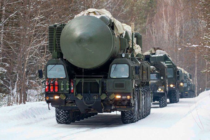 First image shows an older man with white hair and mustache wearing a dark suit white shirt and blue tie sitting in an ornate wooden chair gesturing with his right hand while speaking. Belarusian flags in green red and white are visible behind him. Second image depicts a convoy of large green military vehicles with covered cylindrical payloads resembling missile launchers driving on a snowy road through a forested area in winter with trees and snow-covered ground.