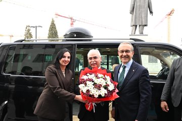 First image shows three people standing beside a black van: a woman in a brown blazer holding a bouquet of red and white flowers extending it to an older man in a suit with a red ribbon, while another man in a blue suit and green tie stands nearby; a statue of a figure in a long coat is visible in the background with construction cranes and trees. Second image depicts two men in suits standing behind a wooden podium in a room with gold-framed walls and multiple Turkish flags on stands, with text reading MALATYA VALİLİĞİ above. Third image features three men in suits posing in front of a similar backdrop with Turkish flags and the MALATYA VALİLİĞİ text on a gold frame. Fourth image shows a group of men in suits seated around a wooden desk in an ornate office with chandeliers, portraits of figures on the wall including one with a red background, Turkish and white flags, and vases of flowers.