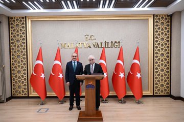 First image shows three people standing beside a black van: a woman in a brown blazer holding a bouquet of red and white flowers extending it to an older man in a suit with a red ribbon, while another man in a blue suit and green tie stands nearby; a statue of a figure in a long coat is visible in the background with construction cranes and trees. Second image depicts two men in suits standing behind a wooden podium in a room with gold-framed walls and multiple Turkish flags on stands, with text reading MALATYA VALİLİĞİ above. Third image features three men in suits posing in front of a similar backdrop with Turkish flags and the MALATYA VALİLİĞİ text on a gold frame. Fourth image shows a group of men in suits seated around a wooden desk in an ornate office with chandeliers, portraits of figures on the wall including one with a red background, Turkish and white flags, and vases of flowers.