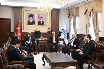 First image shows three people standing beside a black van: a woman in a brown blazer holding a bouquet of red and white flowers extending it to an older man in a suit with a red ribbon, while another man in a blue suit and green tie stands nearby; a statue of a figure in a long coat is visible in the background with construction cranes and trees. Second image depicts two men in suits standing behind a wooden podium in a room with gold-framed walls and multiple Turkish flags on stands, with text reading MALATYA VALİLİĞİ above. Third image features three men in suits posing in front of a similar backdrop with Turkish flags and the MALATYA VALİLİĞİ text on a gold frame. Fourth image shows a group of men in suits seated around a wooden desk in an ornate office with chandeliers, portraits of figures on the wall including one with a red background, Turkish and white flags, and vases of flowers.