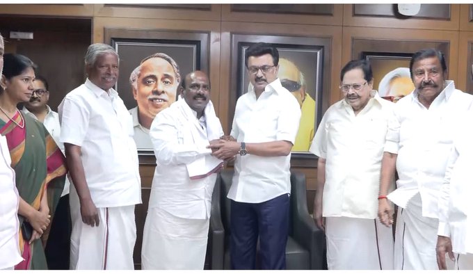 Group of Indian politicians in formal attire including white shirts and dhotis gathered in an office setting with wooden panels and portraits on walls one man in blue pants shaking hands with another while others stand around including women in saris and men with mustaches the central figures appear to be Manoj Pandian and the Speaker
