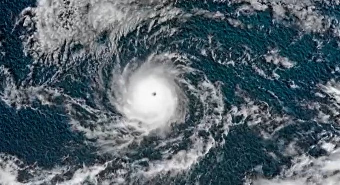 Satellite image displays a large swirling typhoon over the ocean with a prominent white eye at the center surrounded by dense spiral cloud bands in white and gray tones against dark blue waters.