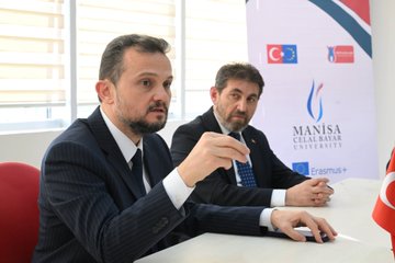 First image shows two men in suits seated at a table in a room with windows and banners for Manisa Celal Bayar University, Erasmus+, and Turkish and EU flags, one man holding a pen. Second image depicts a group of five people including men and women in business attire sitting around a table with pastries, glasses, and notebooks, with posters on walls. Third image features four women in professional clothing seated at a long table with country flags like China and Spain on walls. Fourth image is a large group photo of about 15 men and women in formal attire standing in a hallway with university signage and doors.