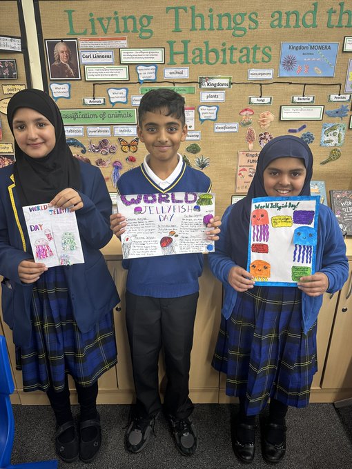 Three children in blue school uniforms stand in front of a classroom display board titled Living Things and Habitats with images of Charles Darwin plants and animals including butterflies and jellyfish drawings. The girl on the left wears a headscarf and holds a book titled World with a jellyfish drawing. The boy in the center holds a poster about jellyfish facts. The girl on the right wears a headscarf and holds a blue paper with an octopus drawing. Wooden shelves and educational posters are visible in the background.