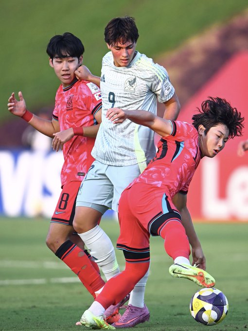 Three soccer players in action on a green field during a match one in red jersey with number 9 from Mexico team raising arm another in gray and green striped jersey from South Korea team extending arm and third in red jersey with number 8 from Mexico team kicking a yellow soccer ball near the ground with red stadium seating and advertising boards visible in the background.