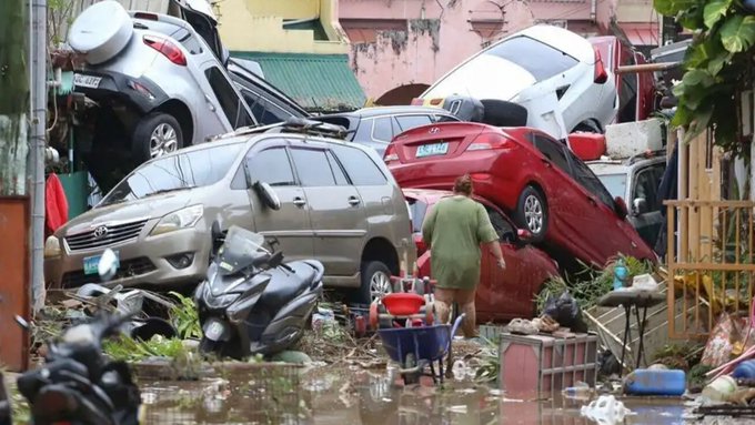 Scene of severe flooding and destruction from typhoon with multiple damaged vehicles including cars and a motorcycle piled up and partially submerged in muddy water. A person in green clothing walks through the debris near overturned cars and scattered household items like buckets and plants. Background shows pink and green buildings with exposed structures amid the floodwaters and wreckage.