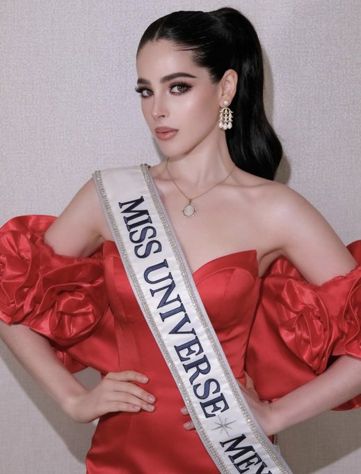 A woman with long dark hair in a ponytail stands posing with hands on hips. She wears a red off-shoulder gown with large puffed sleeves and a fitted bodice. A white sash with gold lettering reading Miss Universe Mexico drapes across her torso. She has gold hoop earrings and a necklace. The background is plain light gray.
