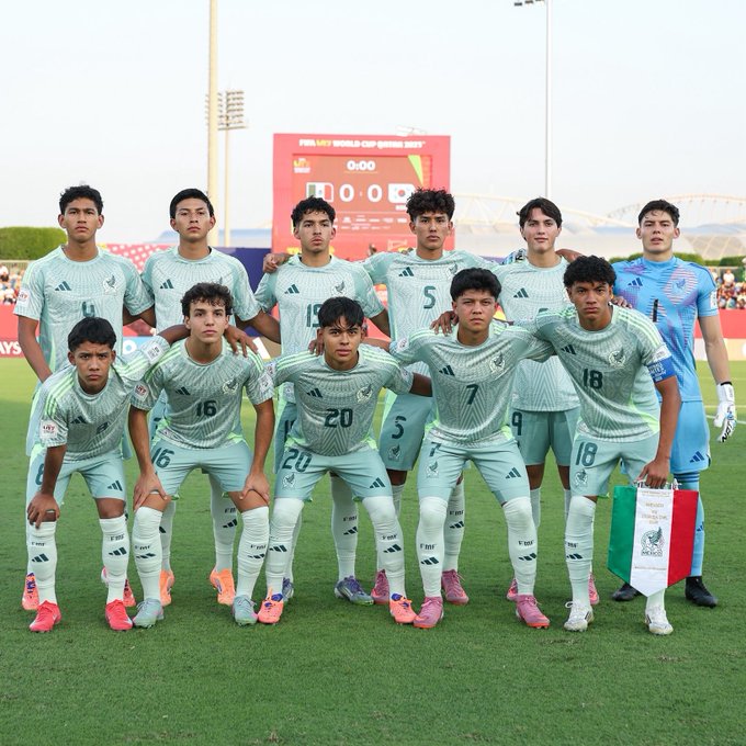 Group of young male soccer players in green and white uniforms with numbers like 16, 5, 18, 20, and 7 stand together on a green field under floodlights, posing with arms around shoulders and hands on knees, in front of a scoreboard showing 0-0 and a banner reading FIFA U-17 WORLD CUP QATAR, with a Mexican flag held by one player.