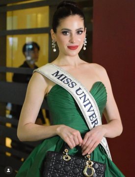 A woman with dark hair in an updo wears large hoop earrings and red lipstick, standing in front of a glass door holding a black handbag, dressed in a strapless green gown with a wide white Miss Universo sash across her chest. A man with short dark hair smiles wearing a white shirt under a dark blazer with a star emblem and MJU label, against a purple background.