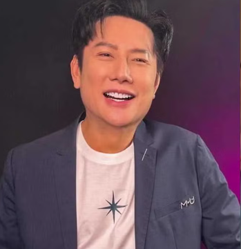 A woman with dark hair in an updo wears large hoop earrings and red lipstick, standing in front of a glass door holding a black handbag, dressed in a strapless green gown with a wide white Miss Universo sash across her chest. A man with short dark hair smiles wearing a white shirt under a dark blazer with a star emblem and MJU label, against a purple background.