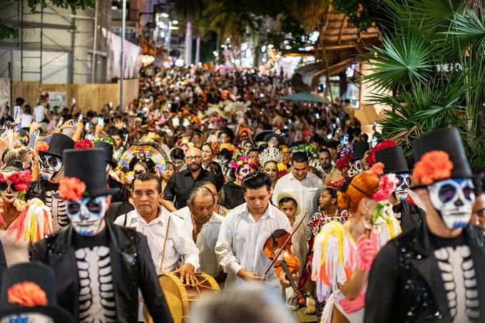 Vibrant nighttime street parade scene with large crowd of participants wearing colorful Day of the Dead costumes including skull masks, top hats with flowers, traditional dresses, and face paint. Musicians play violin and drums while dressed in white shirts and black skeletal outfits. Palm trees and urban buildings line the background under illuminated lights.