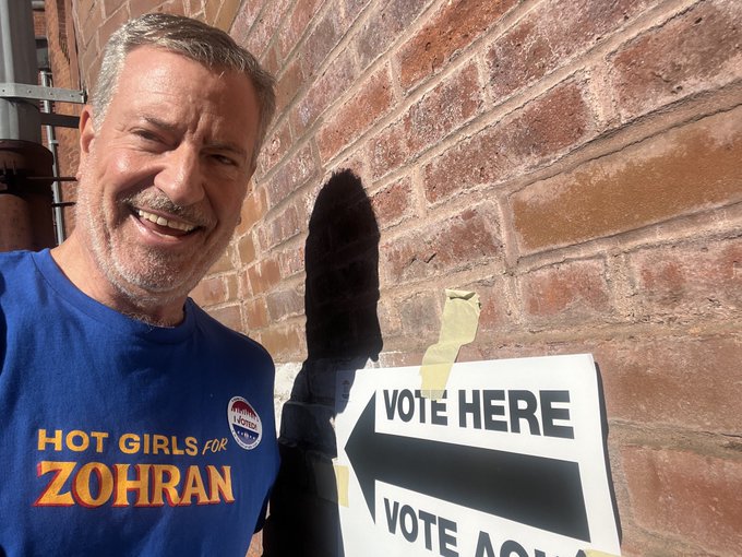 Bill De Blasio Hot Girl For Zohran Shirt 1 A middle-aged man with gray hair and a mustache smiles at the camera while standing outdoors against a red brick wall. He wears a blue t-shirt printed with Hot Girls for Zohran in orange letters and a round blue campaign button on his chest. He holds a white rectangular sign taped to the wall that reads Vote Here in black letters with a black arrow pointing right. A shadow falls across the lower part of the image near the sign.