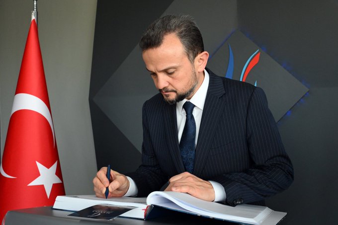 First image shows a man in a dark pinstripe suit and white shirt signing a large open book with a pen at a desk, Turkish flags on either side, geometric wall backdrop. Second image depicts a man in a dark suit and a woman in a green coat and headscarf standing at a podium labeled Manisa Celal Bayar Universitesi, Turkish and white flags nearby, geometric backdrop. Third image features a group of eight formally dressed individuals, including men in suits and a woman in a green coat and headscarf, standing together at a podium with university logo, Turkish and white flags, geometric wall design.