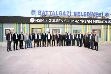 First image shows a large group of men in suits standing in front of a building facade with Battalgazi Belediyesi sign and Asım Gülsen Solmaz Yaşam Merkezi text in colorful striped design. Second image depicts the same group posing near concrete mixing plant equipment with silos and conveyor belts against a hilly landscape. Third image features a smaller group of men including one holding a booklet with landscape image standing on dirt ground with city buildings and construction cranes in background. Fourth image displays men in formal attire gathered at night under string lights and archway structure on paved area.