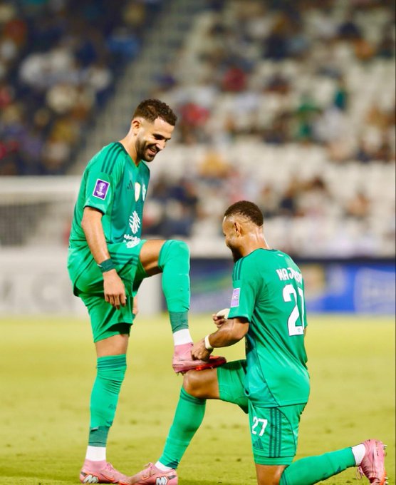 Two male soccer players in green jerseys and shorts stand on a green field with a blurred stadium crowd in the background. One player with short dark hair kneels on his right knee adjusting pink and white cleats on his left foot while smiling. The other player with short hair stands on his left leg lifting his right leg extended behind with knee bent wearing matching pink and white cleats. Both wear green socks pulled up to knees and the kneeling player has number 27 on his jersey while the standing one has number 21. The scene appears post-match celebratory.