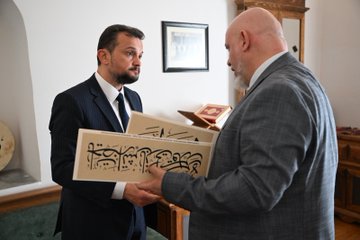 First image shows four people standing outdoors near trees and display boards in a garden setting with fences and a glass structure; two men in suits, one bald with beard in gray suit, one younger in dark suit, and a woman in black with headscarf. Second image depicts a man in white Sufi robe and brown turban raising arms in a room with wooden door, another man in dark suit looking on. Third image features a man in dark suit sitting on green couch in room with wooden furniture, Arabic calligraphy drum on wall, Quran stand with book. Fourth image shows two men in suits, one holding large wooden plaque with Arabic script, standing in room with bookshelves and artwork.