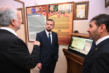 First image shows two men in suits, one with gray hair gesturing while speaking to another with short dark hair, standing in a room with a large map of Europe and Asia on the wall, framed pictures, an arched wooden door, plants, and spotlights. Second image depicts four people including two men in suits, one older with gray hair, and a woman in a headscarf and black clothing, gathered around a wooden table in a library-like room with bookshelves, framed artworks, a TV screen, and Arabic script on walls. Third image features three men in suits, one with gray hair, one with a mustache, and one younger, standing near wall displays with text panels, artworks, and a digital screen in a ornate room with wooden furniture. Fourth image shows a group of six people in formal attire, including men in suits and two women in headscarves and abayas, posing together outdoors near an arched stone entrance with decorative wall art and wooden furniture in a courtyard setting.