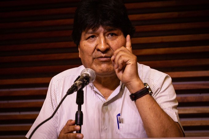 First image shows Evo Morales, a man with dark hair and indigenous features, standing at a wooden podium with a microphone, wearing a light shirt and watch, gesturing with his right hand index finger extended while speaking in an indoor setting with wooden paneling behind. Second image depicts Nicolas Maduro, a man with gray hair and mustache, smiling and clapping with both hands, dressed in a dark suit and gold chain, wearing a yellow, blue, and red sash with stars representing the Venezuelan flag, standing in an indoor venue with a blurred background.