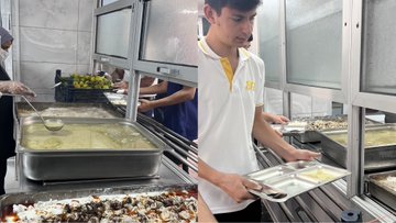 First image shows two educational posters one titled Yeterli ve Dengeli Beslenme with illustrations of milk cheese bread fruits vegetables and grains emphasizing adequate balanced nutrition in child development the other titled Yiyecekleri Okuldan Alıyorum Sağlıklı Besleniyorum with a pyramid of food groups including grains vegetables fruits proteins dairy and fats plus text on balanced plate composition. Second image depicts stacked yogurt containers on shelves in a storage area with a light bulb overhead and three individuals wearing hairnets and uniforms one holding a bowl of food in a kitchen with stainless steel counters refrigerator and ventilation. Third image shows a person holding a package of cereal labeled with red branding another wearing a hairnet examining items near a freezer with stacked boxes and three men in uniforms one with glasses inspecting inside an old-fashioned refrigerator. Fourth image illustrates people in hairnets and uniforms handling food in a serving area one scooping from a large pot of white substance another placing items on trays with stainless steel surfaces baskets of green grapes trays of prepared food like rice and vegetables behind a serving counter.