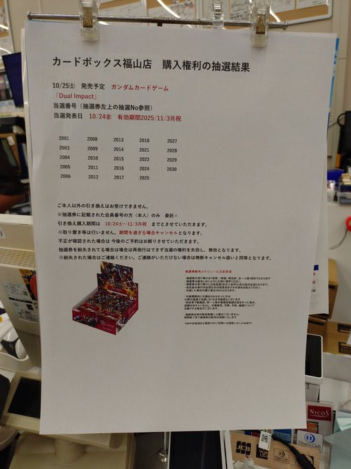 Poster on wall with text announcing winning numbers for Gundam Card Game Dual Impact lottery listing numbers from 2000 to 2024 in a table format below that additional Japanese text explaining rules and requirements stacks of card game boxes on shelves in background including colorful Gundam themed packages and a Nacos branded item