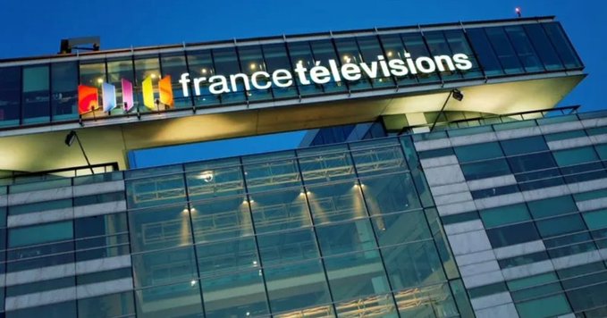 Modern glass building at dusk with illuminated colorful logo reading France Télévisions on the facade including multicolored stripes above the text on a bridge-like upper structure surrounded by blue sky and lit windows.