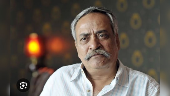 Elderly Indian man with gray hair, mustache, and expressive eyes, dressed in a white shirt, seated against a dark textured wall with circular patterns, a lit orange lamp visible to the right, and a blurred indoor setting in the background.
