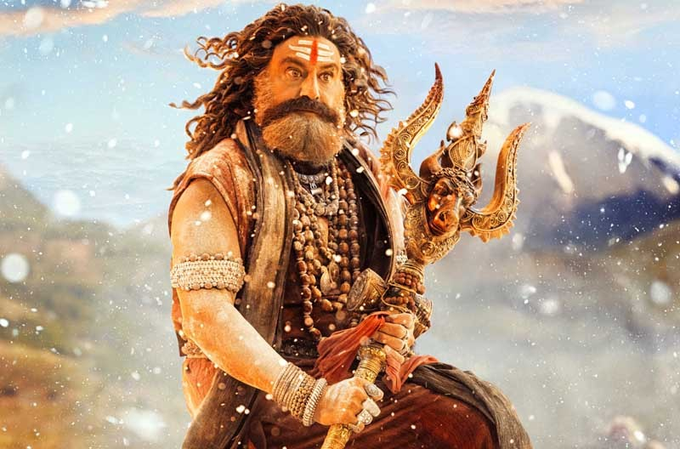 Balakrishna portrayed as a fierce Aghori character with long curly hair, thick beard, red tilak on forehead, wearing saffron upper garment and lower wrap, multiple bead necklaces, armbands, holding ornate trident, standing dynamically against snowy mountain background with mist.