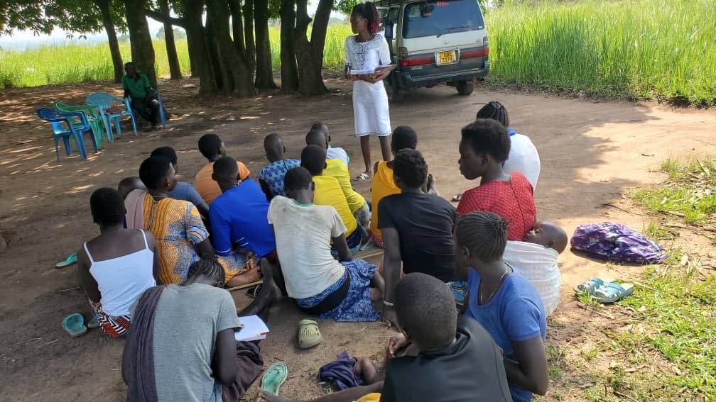 A diverse group of about 15-20 Black African adolescents and young adults mostly men and women in casual colorful clothing like t-shirts dresses and pants sit on the ground or plastic chairs in a circle on a dirt path under shady trees near a lakeside grassy area with a white van parked nearby. A female facilitator in a white top and skirt stands addressing the group gesturing with her hands while some participants hold notebooks and appear engaged in discussion. The setting is outdoors in a rural community environment with green vegetation and water in the background.