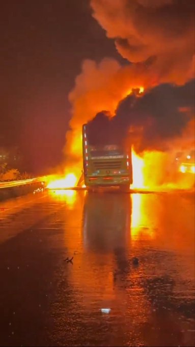 A large bus engulfed in intense orange and yellow flames with thick black smoke billowing upwards captured at night on a wet road surface reflecting the fire light. The bus appears damaged at the rear where the fire originates near the fuel tank area following a collision. Surrounding environment includes a highway with visible lane markings and distant lights from vehicles.