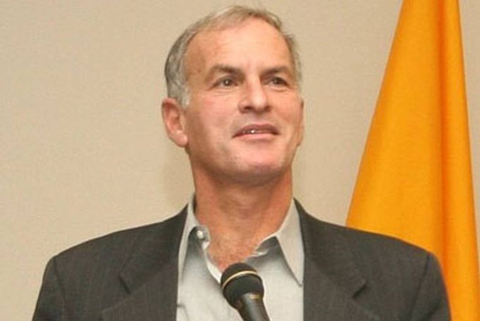 An older man with gray hair and light skin wearing a dark suit and white shirt speaks into a black microphone at a podium in front of a plain background with an orange flag visible to his right. A second image displays the Palestinian flag featuring black, white, green, and red triangular and rectangular sections.