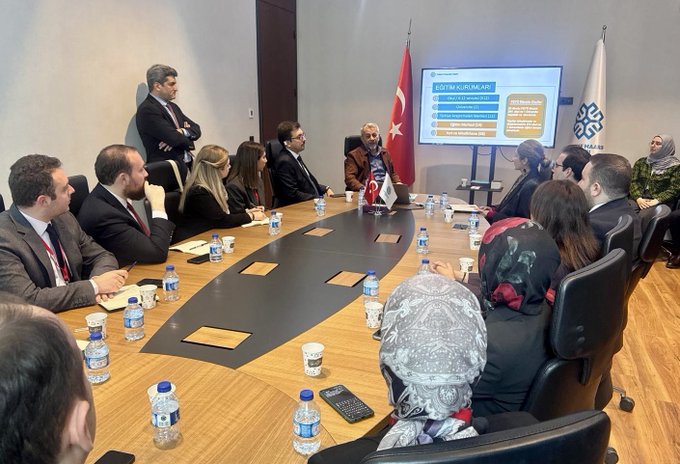 Birinci görüntüde resmi giyimli bir grup kişi oval bir masanın etrafında oturuyor, masada su şişeleri, belgeler ve fincanlar var. Masanın ucunda mavi ekran üzerinde sarı ve kırmızı grafikli sunum gösteriliyor, yanında Türk bayrağı ve beyaz zeminli mavi sembol bayrağı asılı. Arka planda ayakta duran gri takım elbiseli bir adam kollarını kavuşturmuş bakıyor, oda ahşap zeminli ve modern mobilyalı. İkinci görüntüde çeşitli milletlerden resmi giyimli bir grup kişi ayakta poz veriyor, arka planda koyu mavi duvar üzerinde mavi sembol, Türk bayrağı ve başka bayraklar asılı.