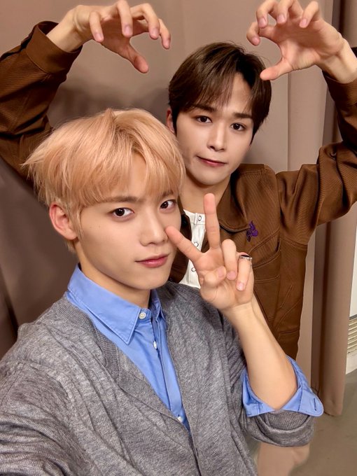 Two young men stand side by side in a studio setting with a beige curtain background. The one on the left has light pink hair, wears a blue shirt under a gray jacket, and makes a peace sign with his right hand while smiling. The one on the right has brown hair, wears a brown jacket over a white shirt, and forms claw shapes with both hands while smiling. Kimura Masaya and Gotoh Takanori from INI pose playfully.