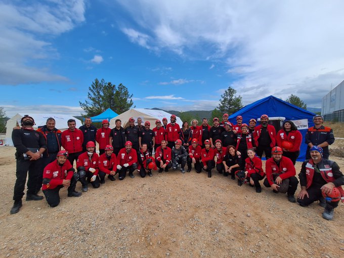 First image shows a large group of personnel in red uniforms and helmets posing together outdoors near tents and trees under a clear sky, including men and women kneeling and standing in formation on dirt ground. Second image depicts four rescuers in red gear and helmets performing a training drill, carrying a person on a stretcher from a simulated structure made of plywood walls with graffiti and windows, set in an outdoor area with trees and blue sky.