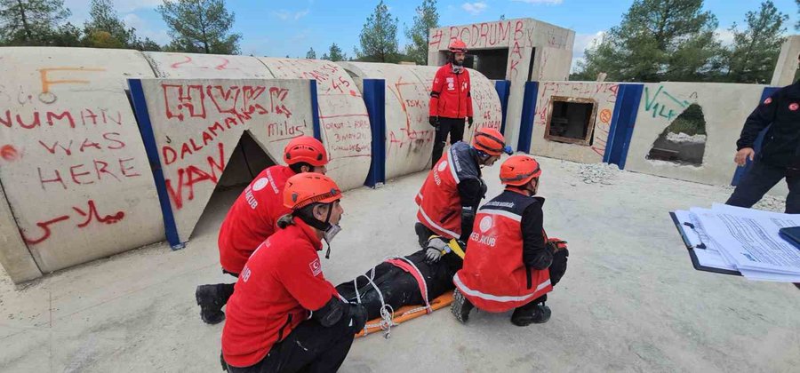 First image shows a large group of personnel in red uniforms and helmets posing together outdoors near tents and trees under a clear sky, including men and women kneeling and standing in formation on dirt ground. Second image depicts four rescuers in red gear and helmets performing a training drill, carrying a person on a stretcher from a simulated structure made of plywood walls with graffiti and windows, set in an outdoor area with trees and blue sky.