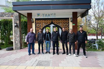 First image shows five men including police officers in uniforms sitting around a metal table outdoors on grass near parked cars and trees, engaged in conversation with one holding a cup. Second image depicts eight men standing in front of a building entrance with TAKSIT sign and decorative arches, some in police attire, posed for a group photo on a paved area with plants. Third image features three men seated on chairs near a wooden bench outside a brick building, two in police vests discussing. Fourth image captures two men shaking hands inside a jewelry store filled with gold displays and necklaces on counters, one in police uniform and the other in casual shirt.