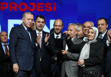 First image shows a man in a dark suit standing at a white podium holding a microphone with his right hand raised gesturing, speaking into it, behind him a large green and orange illuminated backdrop with Turkish text reading YÜZYIL KONUT PROJESİ and 500 BİN SOSYAL KONUT in bold letters, stage lights and a red emblem on the podium. Second image depicts the same man in a suit at a podium with a microphone, facing a large audience seated in a spacious indoor venue with green plants and flowers around the stage, multiple blue screens displaying EVSAHIBI and other text, bright overhead lights and a screen showing the mans face. Third image features a group of about ten people mostly in suits including the central man in a blue suit with a red tie clapping, a woman in a headscarf and black outfit next to him, others holding a black tablet device, against a blue backdrop with white text PROJESİ. Fourth image captures a large group of over twenty people standing in a row on a stage, many in suits some women in formal attire, central figures including the man in blue suit, large screens on sides showing the mans face, blue EVSAHIBI text in the center, surrounded by green plants and stage lighting in a grand hall setting.