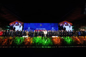 First image shows a man in a dark suit standing at a white podium holding a microphone with his right hand raised gesturing, speaking into it, behind him a large green and orange illuminated backdrop with Turkish text reading YÜZYIL KONUT PROJESİ and 500 BİN SOSYAL KONUT in bold letters, stage lights and a red emblem on the podium. Second image depicts the same man in a suit at a podium with a microphone, facing a large audience seated in a spacious indoor venue with green plants and flowers around the stage, multiple blue screens displaying EVSAHIBI and other text, bright overhead lights and a screen showing the mans face. Third image features a group of about ten people mostly in suits including the central man in a blue suit with a red tie clapping, a woman in a headscarf and black outfit next to him, others holding a black tablet device, against a blue backdrop with white text PROJESİ. Fourth image captures a large group of over twenty people standing in a row on a stage, many in suits some women in formal attire, central figures including the man in blue suit, large screens on sides showing the mans face, blue EVSAHIBI text in the center, surrounded by green plants and stage lighting in a grand hall setting.