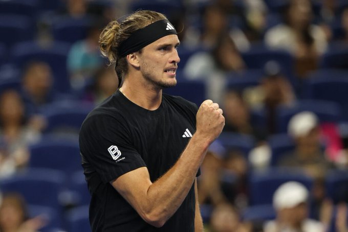 A male tennis player with long blond hair tied in a ponytail wears a black Adidas headband and sleeveless black shirt with Adidas logos on the arms, standing on a blue tennis court surrounded by a crowd in stands, raising his right fist in celebration while looking determined.