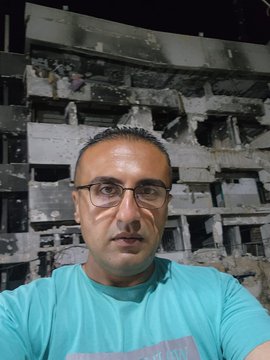 First image shows a man with glasses wearing a blue t-shirt standing outdoors near a white building with Arabic signs indicating a laboratory entrance and door. Second image depicts the same man with glasses in front of a damaged multi-story building at night with debris and people visible. Third image portrays the man with glasses sitting in an office wearing a pink short-sleeve shirt near documents and a computer monitor.