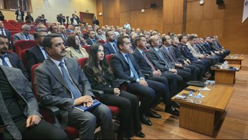 First image shows a conference room with a long white table covered in cloth, featuring a computer monitor, documents, glasses of water, and flower arrangements; three men in suits sit at the table, one with glasses and a mustache, another in a dark suit, and a third in a gray suit; Turkish flags and a banner reading BATTALGAZİ BELEDİYESİ hang in the background against wooden panels and stage curtains. Second image depicts rows of red chairs in an auditorium filled with men and women in formal business attire, including suits and dresses, seated and facing forward; small wooden tables with water bottles are placed in front of some rows; wooden walls and a Turkish flag are visible in the setting. Third image captures additional rows of seated attendees in suits and formal wear in the same auditorium, with some holding notebooks, water bottles on side tables, and the wooden interior with a flag in the background.