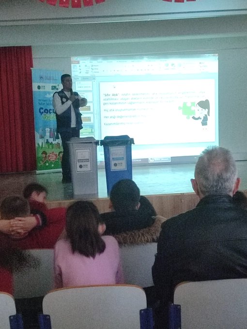 First image shows a classroom with students in red uniforms seated at blue desks, a teacher in a suit pointing at a smartboard displaying content, Turkish flag on wall, educational posters, and children attentively watching. Second image depicts another classroom scene with boys in blue uniforms at orange-trimmed desks, a male teacher in brown jacket gesturing while speaking, world map and flags on walls, backpacks hanging. Third image captures an assembly hall with rows of chairs filled with students in red uniforms, stage with Turkish flags, projector screen showing presentation, adults in suits on stage addressing audience. Fourth image features a stage setup with a man in gray vest speaking at podium, projector displaying text and images related to zero waste including a cartoon character, blue recycling bins nearby, small group of children seated in front rows.