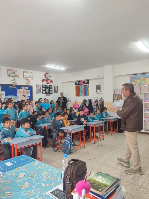 First image shows a classroom with students in red uniforms seated at blue desks, a teacher in a suit pointing at a smartboard displaying content, Turkish flag on wall, educational posters, and children attentively watching. Second image depicts another classroom scene with boys in blue uniforms at orange-trimmed desks, a male teacher in brown jacket gesturing while speaking, world map and flags on walls, backpacks hanging. Third image captures an assembly hall with rows of chairs filled with students in red uniforms, stage with Turkish flags, projector screen showing presentation, adults in suits on stage addressing audience. Fourth image features a stage setup with a man in gray vest speaking at podium, projector displaying text and images related to zero waste including a cartoon character, blue recycling bins nearby, small group of children seated in front rows.