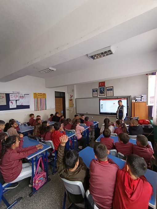 First image shows a classroom with students in red uniforms seated at blue desks, a teacher in a suit pointing at a smartboard displaying content, Turkish flag on wall, educational posters, and children attentively watching. Second image depicts another classroom scene with boys in blue uniforms at orange-trimmed desks, a male teacher in brown jacket gesturing while speaking, world map and flags on walls, backpacks hanging. Third image captures an assembly hall with rows of chairs filled with students in red uniforms, stage with Turkish flags, projector screen showing presentation, adults in suits on stage addressing audience. Fourth image features a stage setup with a man in gray vest speaking at podium, projector displaying text and images related to zero waste including a cartoon character, blue recycling bins nearby, small group of children seated in front rows.